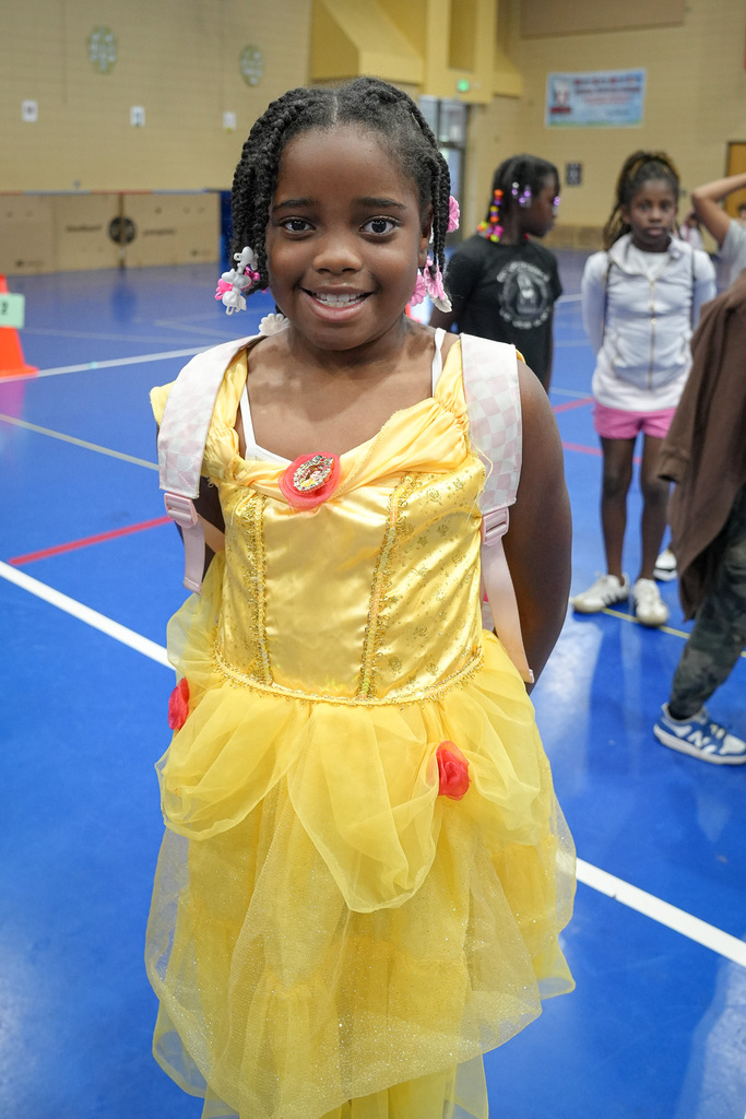 A girl wearing a yellow princess dress stands in a school gym and smiles for a photo.