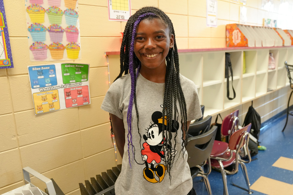 A girl wearing a Mickey Mouse shirt stands inside a classroom and smiles for a photo.