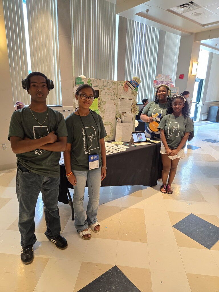 A group of students wearing matching gray shirts with the outline of the state of Alabama on them smile for a photo inside a building. They stand in front of a tri-fold poster board.