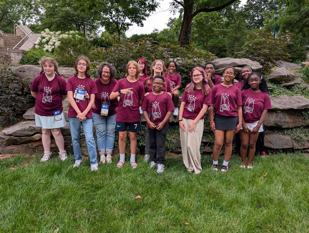 A group of students wearing matching Alabama Future Problem Solvers shirts stand outdoors and smile for a photo. Their coaches stand with them and smile as well.