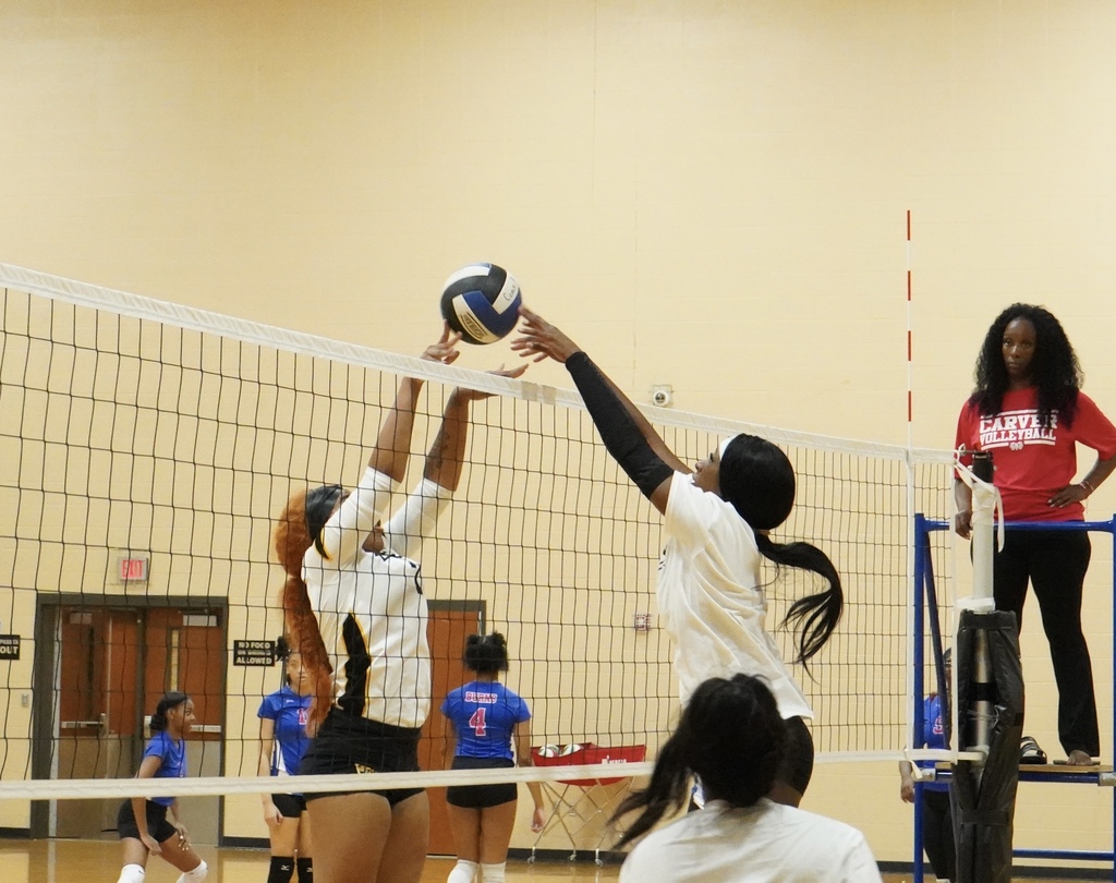 Players from opposing volleyball teams jump toward the net to hit a volleyball.