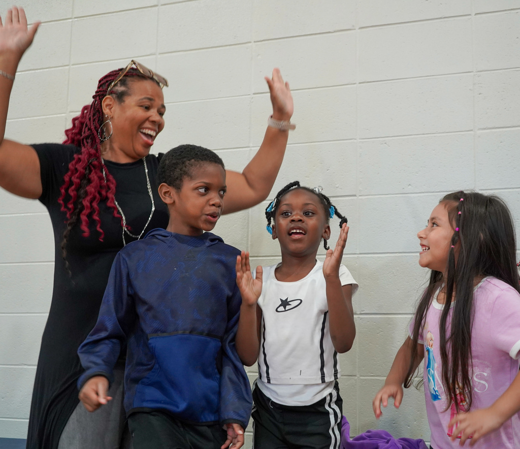 A teacher and students smile and dance during a performance inside a school gym.