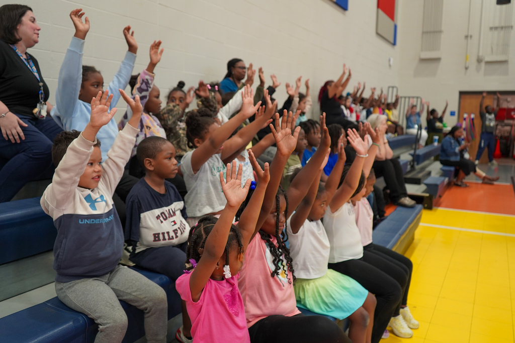 Students hold their hands in the air while sitting in bleachers and watching a performance inside a school gym.