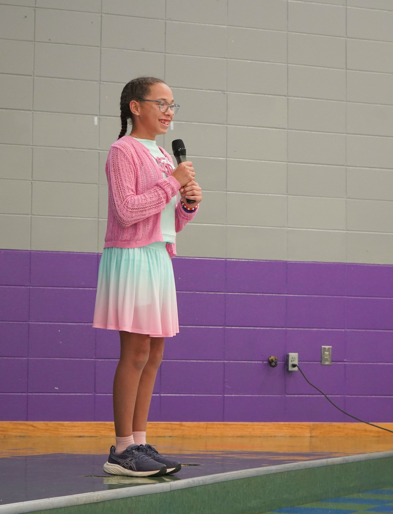 A student smiles and holds a microphone while standing on stage at a school.