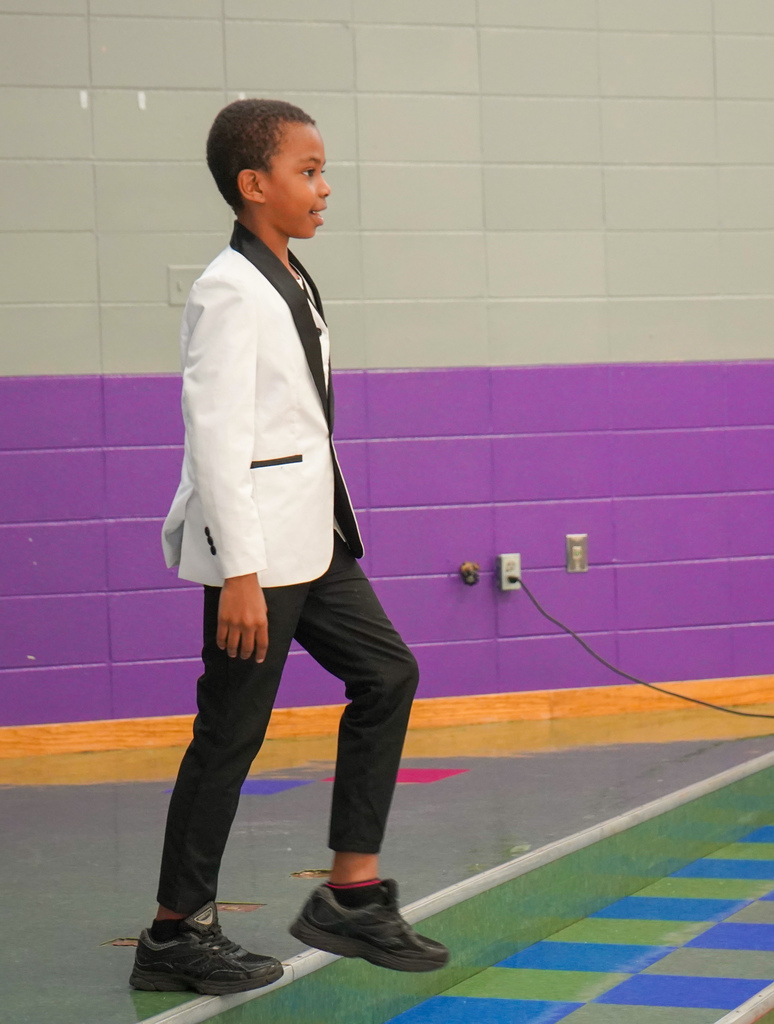 A student wearing a white suit jacket and black pants performs on a stage.