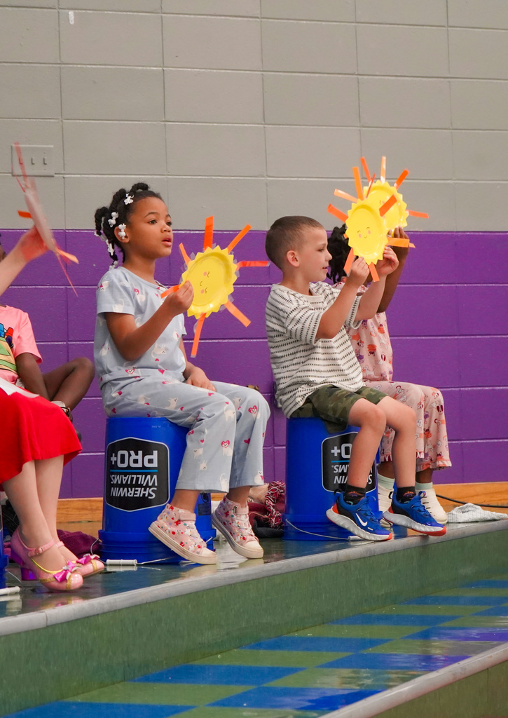 Students perform on stage. They sit on buckets and hold hand-made sunshine art while sitting on buckets.