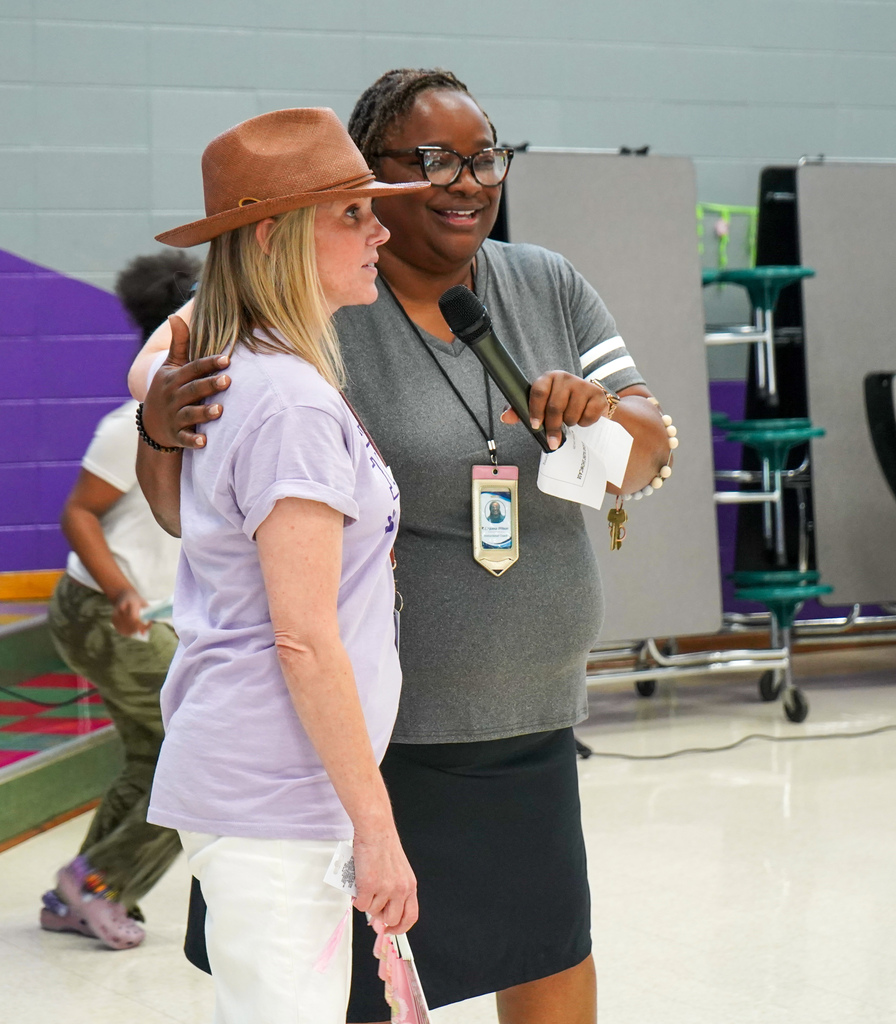 A woman hugs another woman and holds a microphone for her to speak into. They stand inside a school cafeteria.