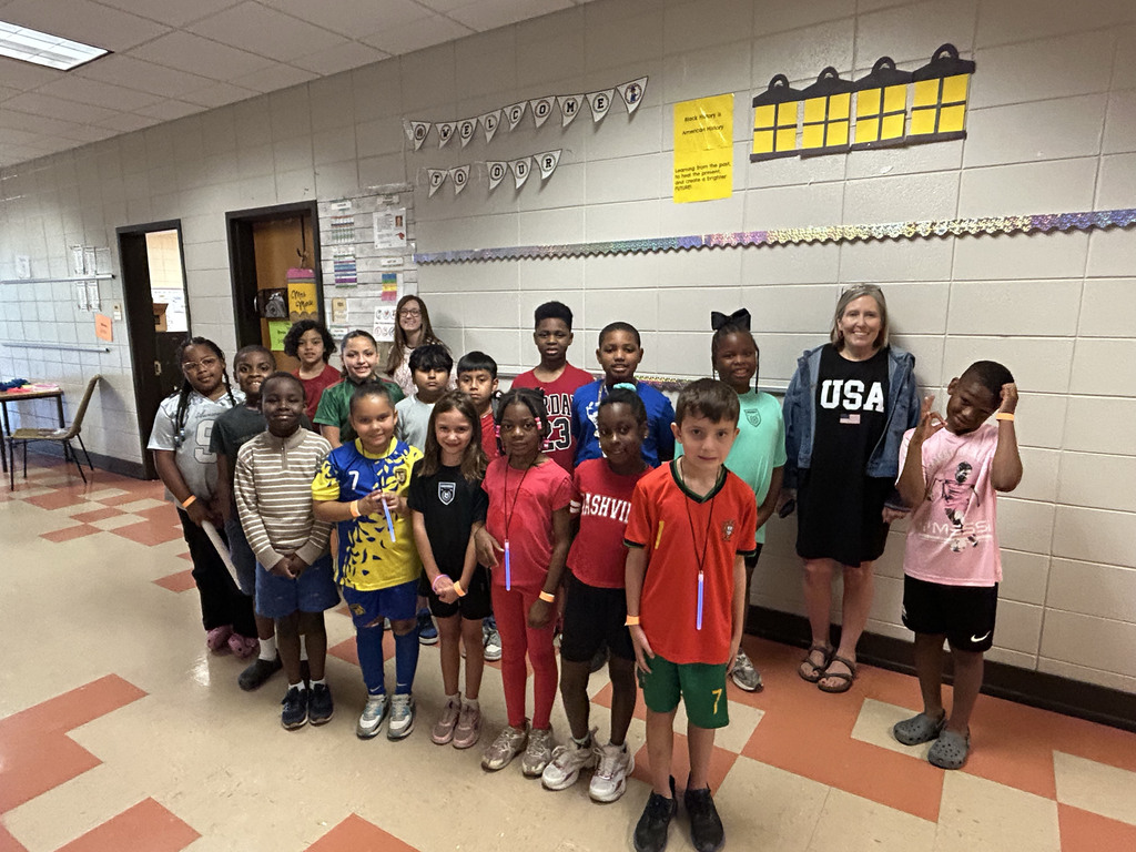 Students and teachers stand in a school hallway and smile for a photo. They all wear outfits supporting different teams playing in the World Cup.