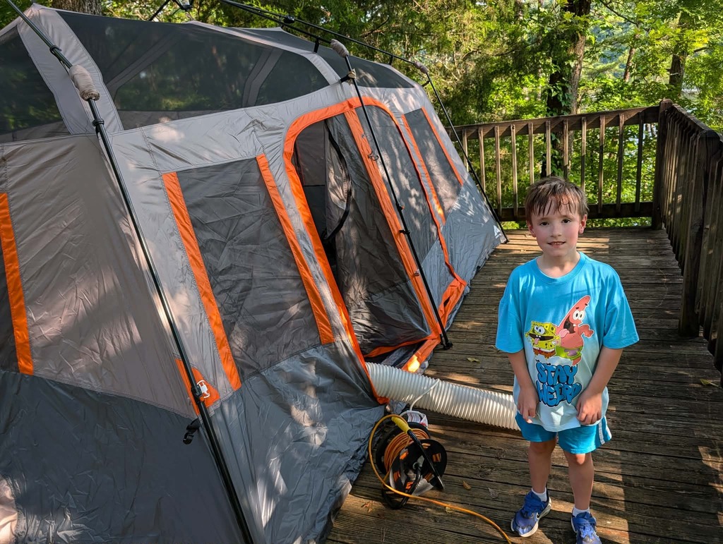 A boy wearing a Spongebob t-shirt smiles while standing next to a tent.