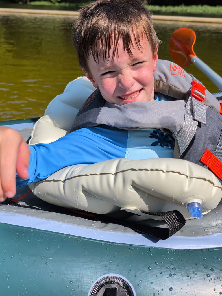 A boy wearing a life jacket smiles while sitting in a kayak on a lake.