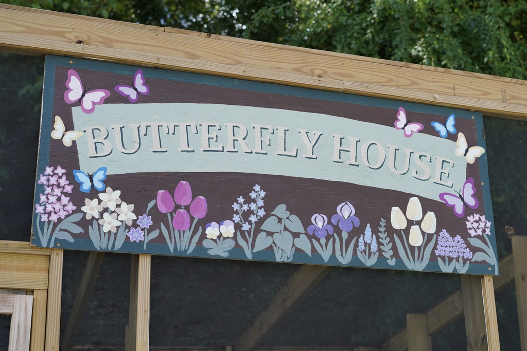 A sign on the outside of a butterfly enclosure says "Butterfly House" and has flowers and butterflies painted on it.