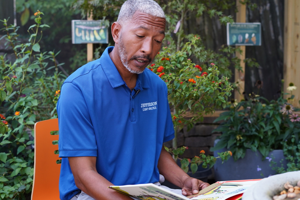 A man sits in a chair surrounded by flowers and reads a book out loud.