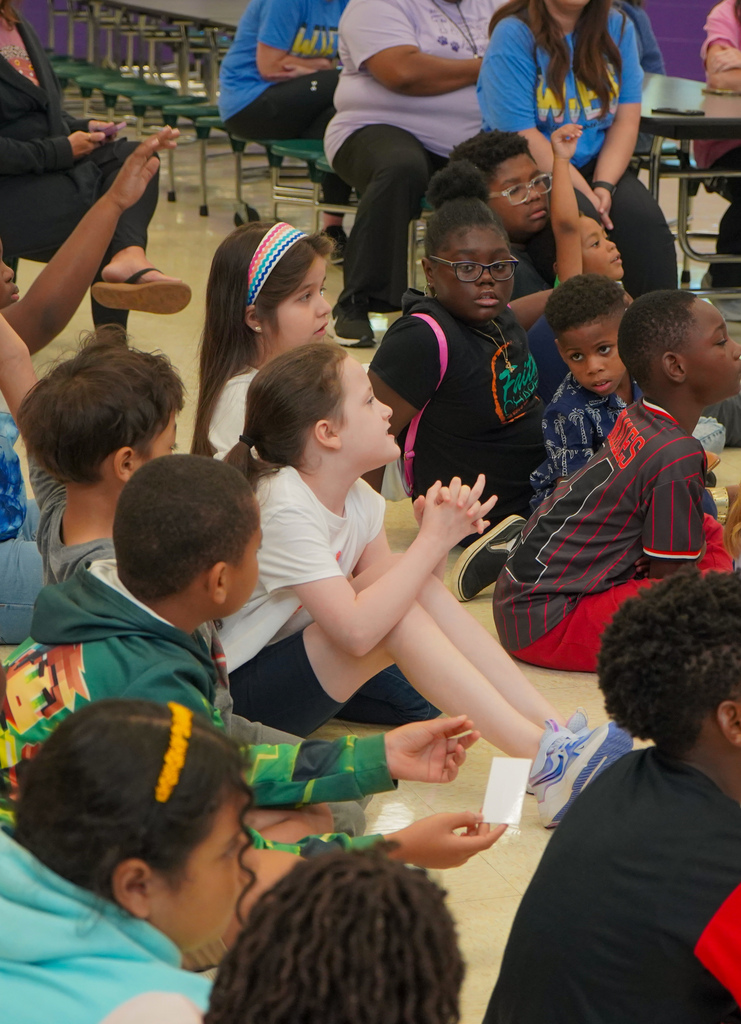 Students sit on the floor of a lunch room. A girl in the middle of the students talks to the group.