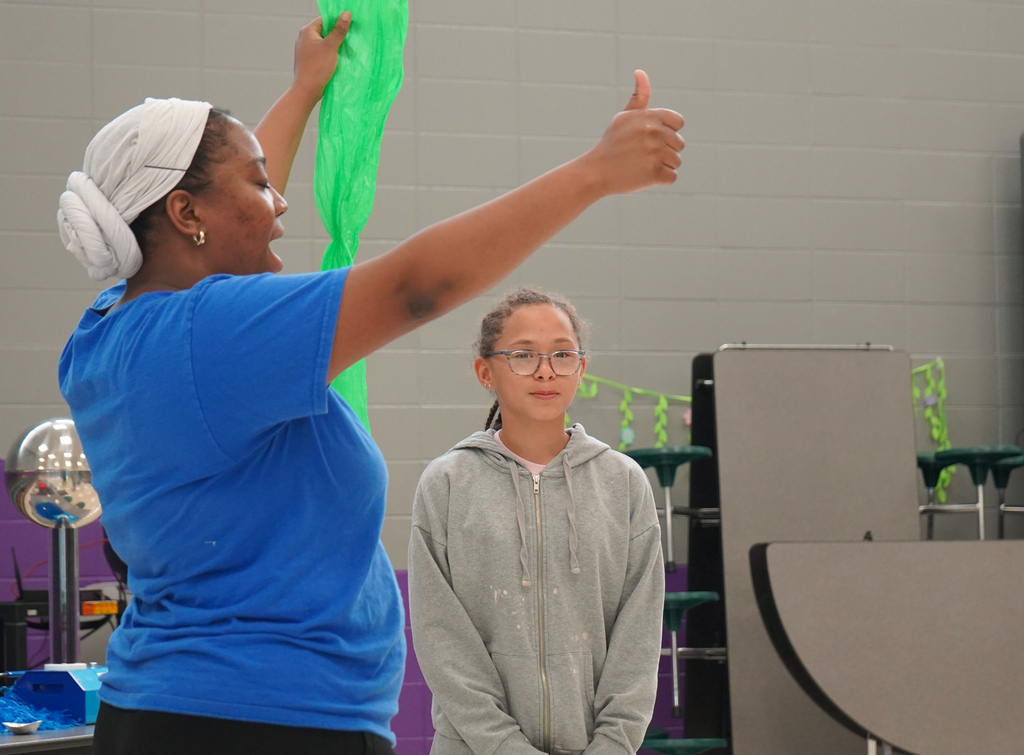 A woman holds a bright green bag in one and hand gives a thumbs up with her other hand. A student stands next to her and watches. They stand inside a school lunch room.