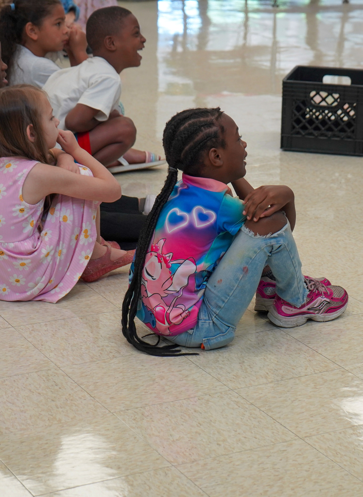 Students smile while sitting on the floor of a lunch room and watching a presentation.