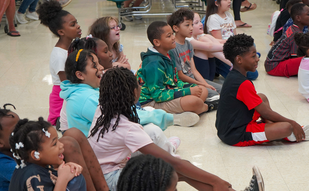 Students smile in awe while sitting on the floor of a lunch room watching a presentation.