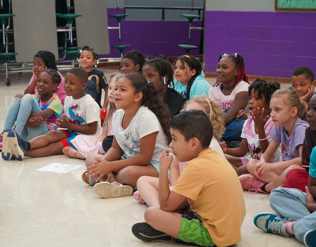 Students sit on the floor of a lunch room and smile while listening to a presentation.
