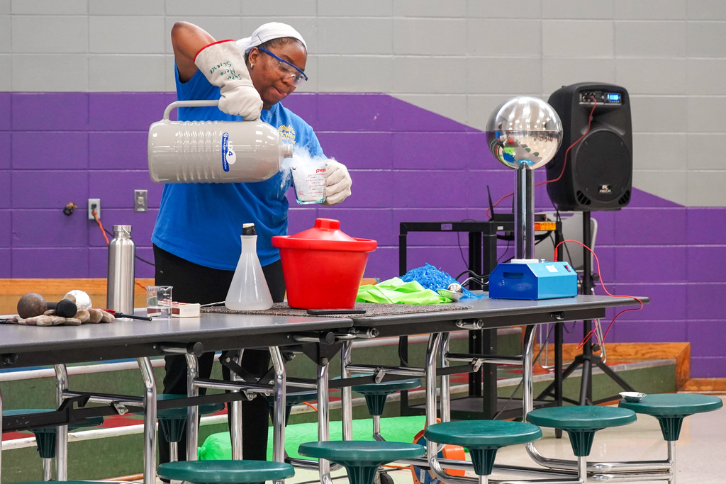 A woman pours a liquid into a measuring cup while performing science experiments in front of children inside a lunch room.