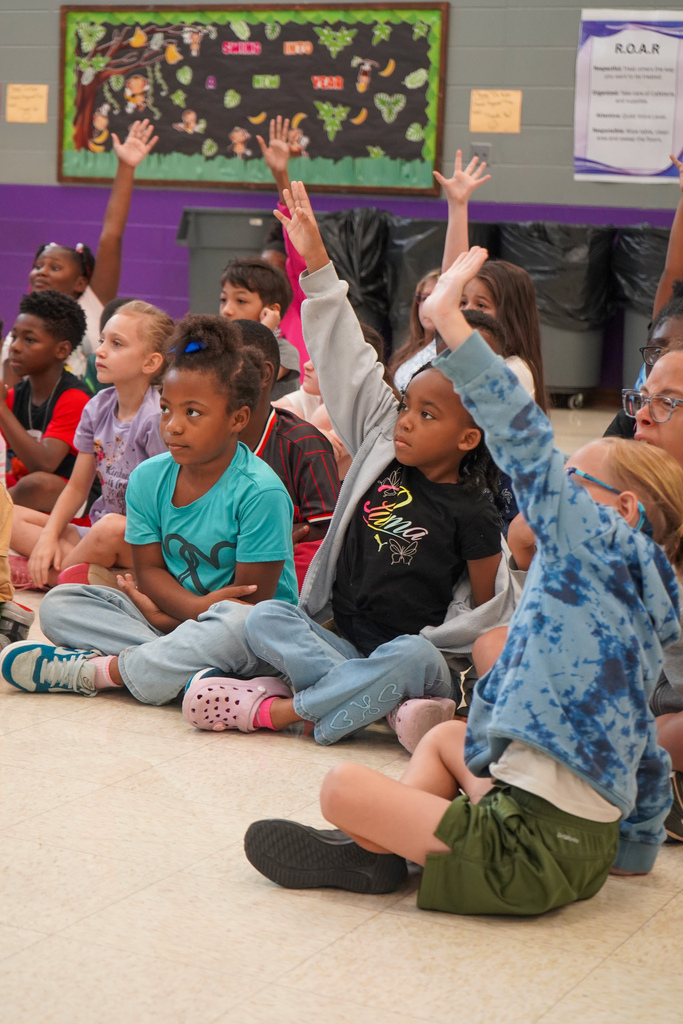 Students raise their hands while sitting on the floor of a lunch room.
