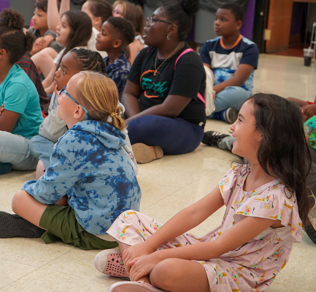 Students smile in awe while watching a presentation. They sit on the floor of a lunch room.