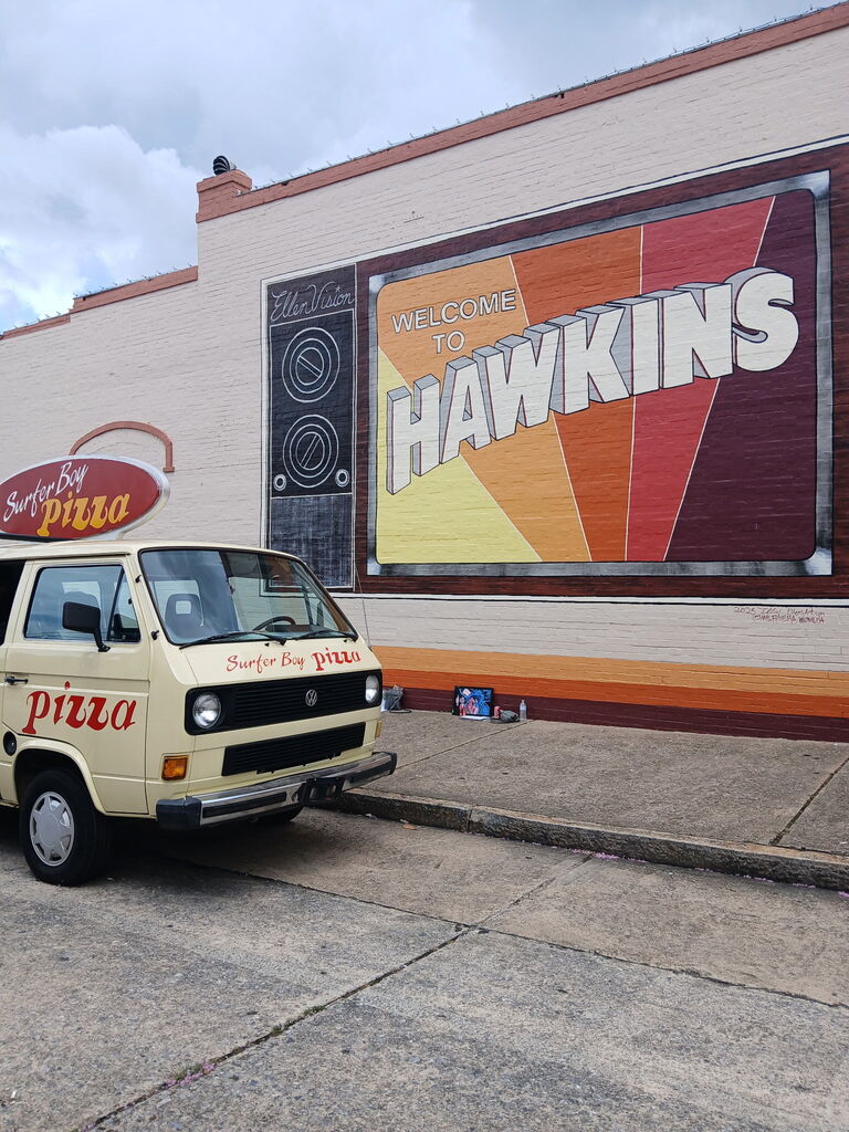 A van with "Surfer Bay Pizza" on the side is parked outside a building with a mural painted on it that says "Welcome to Hawkins."