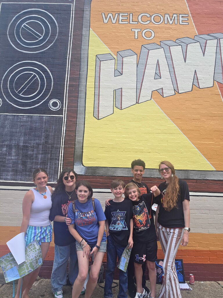 A group of women and students stand together for a photo outdoors. They pose in front of a mural that says "Welcome to Hawkins."