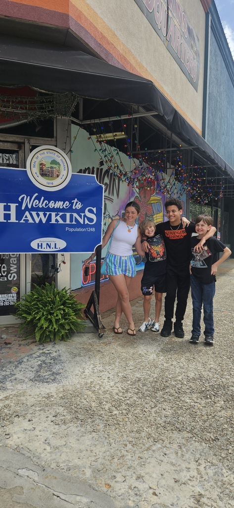 Four people stand together for a photo outside a building. They pose next to a sign that says "Welcome to Hawkins."