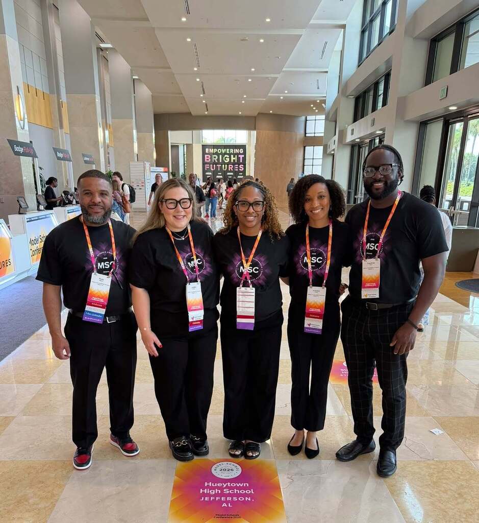 Administrators and teachers from Hueytown High School stand together inside a building and smile. They wear matching Model School Conference t-shirts, and stand on a sign that says Hueytown High School, Jefferson, Alabama.