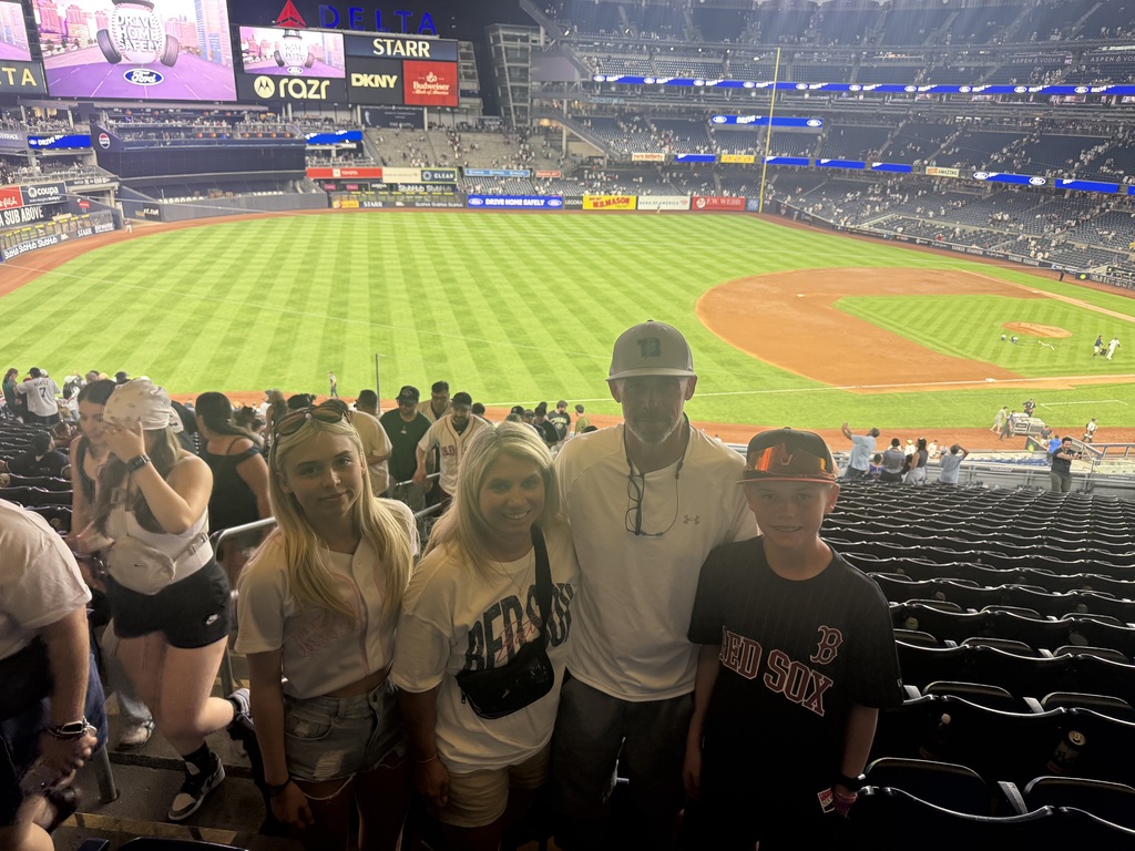 A girl, a woman, a man, and a boy stand together for a photo at a baseball game. They stand in the stadium with the field in the background.