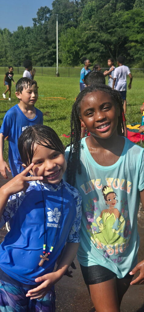 Two girls smile for a photo while standing outdoors. They are completely soaked with water after a water day outside.