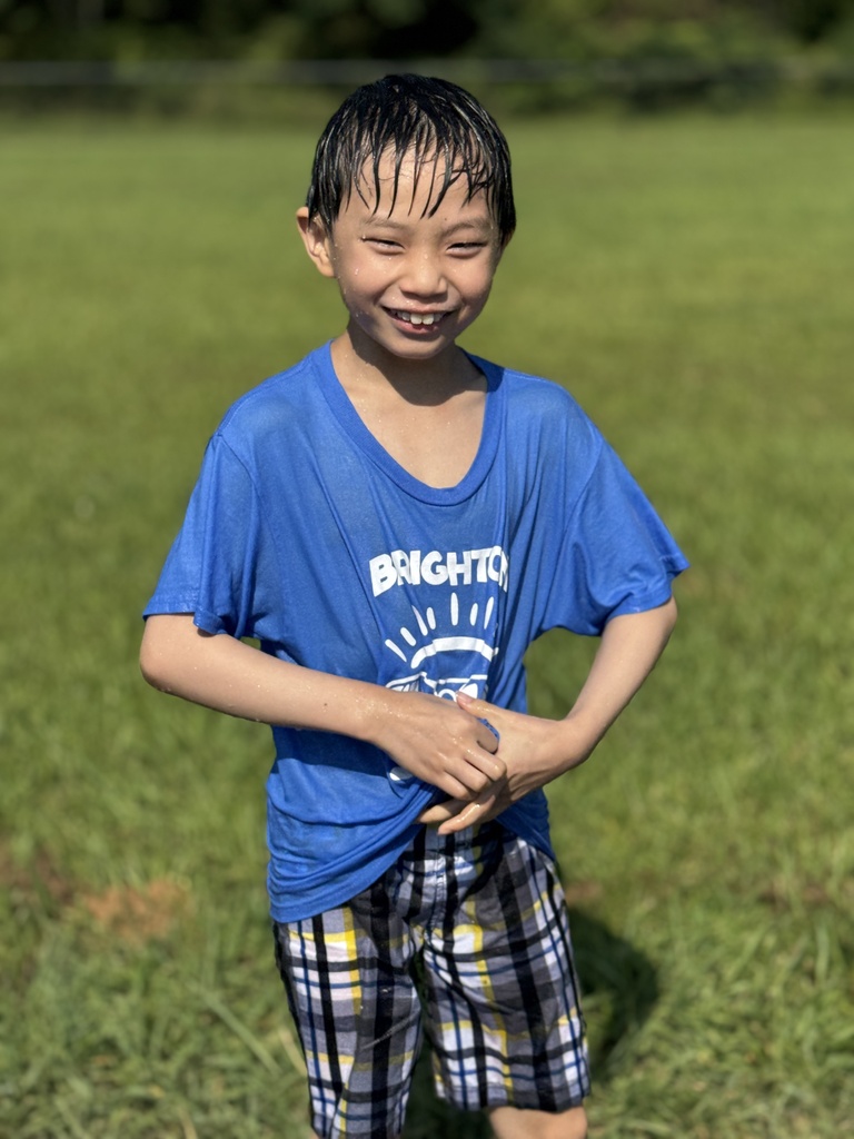 A boy smiles while standing outside on grass. His clothes and hair are soaked with water from a water play day.
