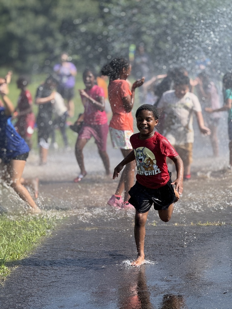 A student smiles while running barefoot through water on pavement. Students play in the falling water behind him.