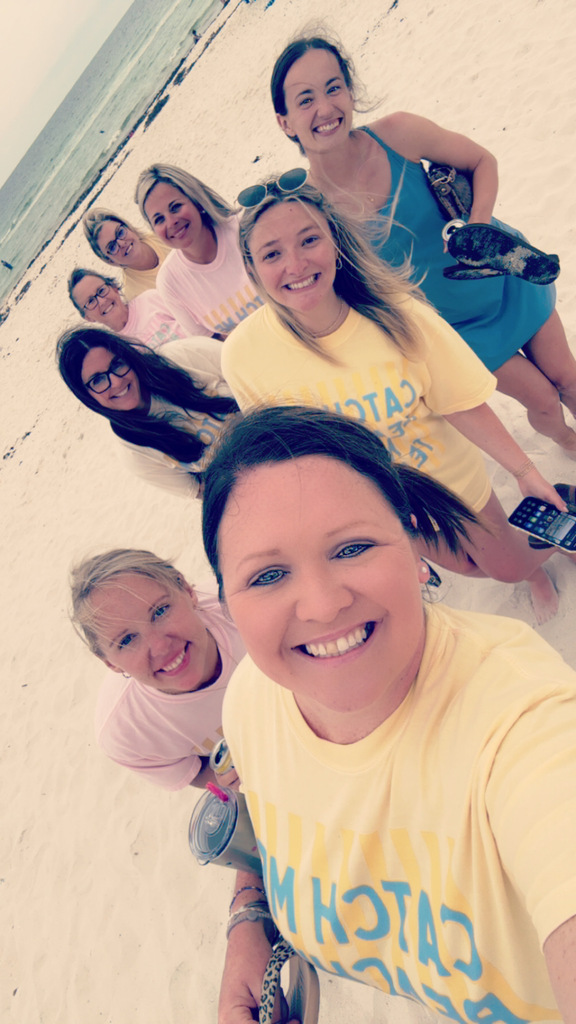 A group of women stand together on a beach and smile for a selfie.
