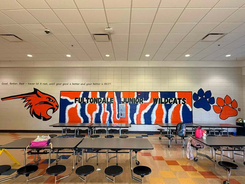 A colorful mural on the wall of the Fultondale Elementary School lunch room. The mural consists of a wildcat, paw prints, and painted words that say "Fultondale Junior Wilcats." Above the mural, a quote is on the wall. It says "Good, better, best — Never let it rest, until your good is better and your better is BEST."