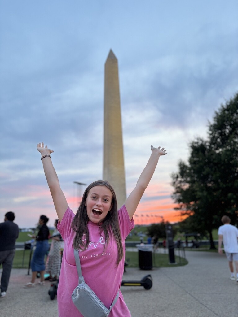 A girl smiles and holds her arms in the air for a photo in front of the Washington Monument during sunset.