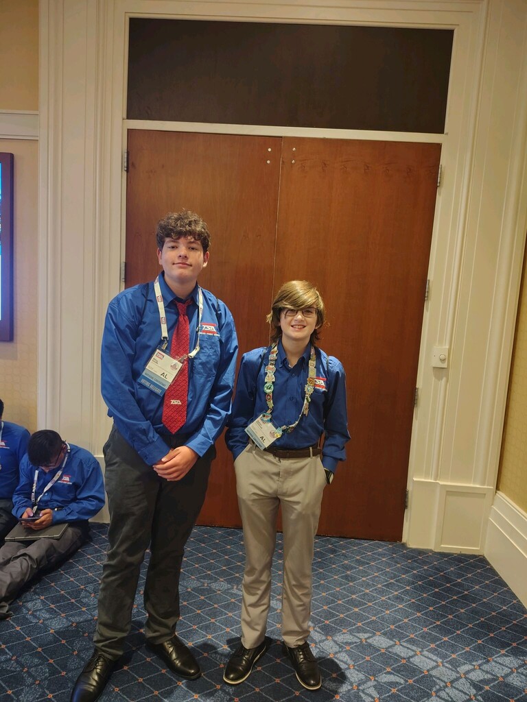 Two boys stand together for a photo. They wear matching blue shirts and lanyards.