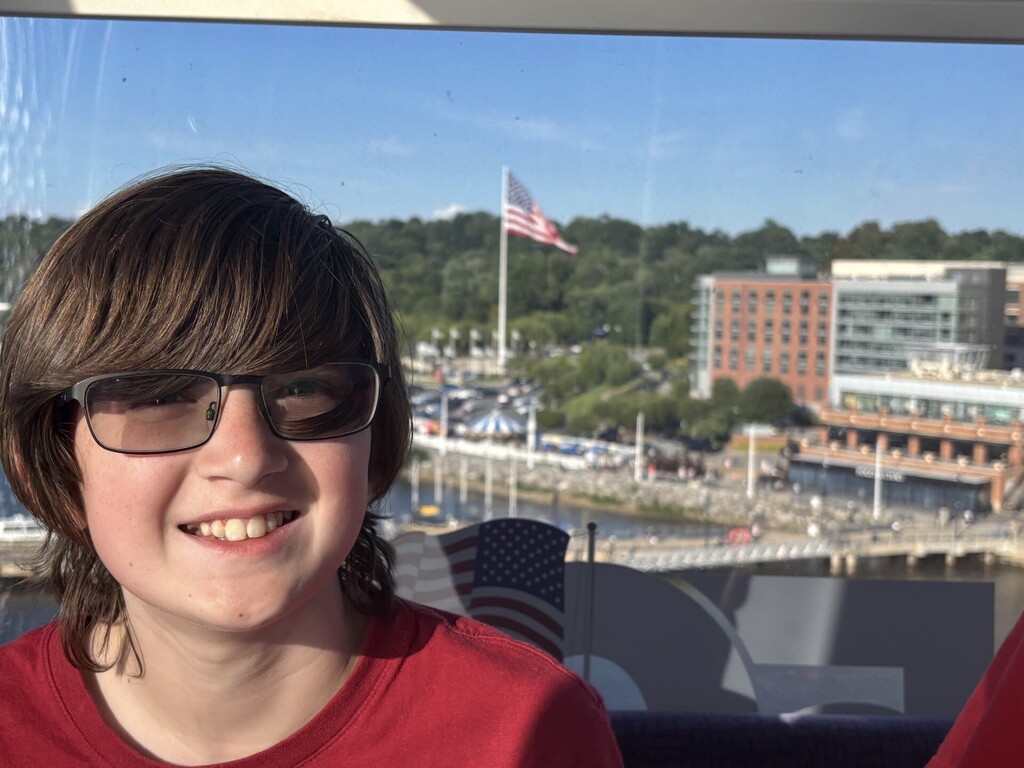 A boy smiles for a photo. Behind him in the background are buildings and an American flag.