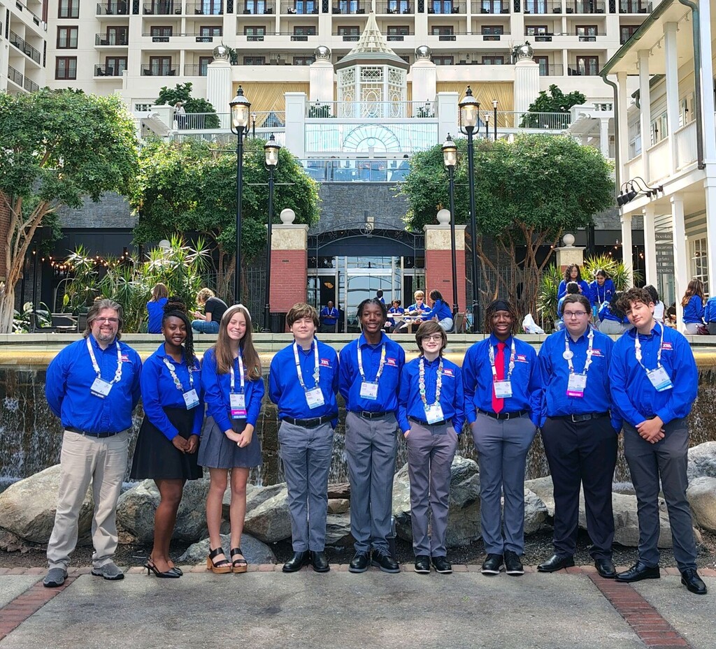 A group of students stand together for a photo in front of a building. They wear matching blue shirts.