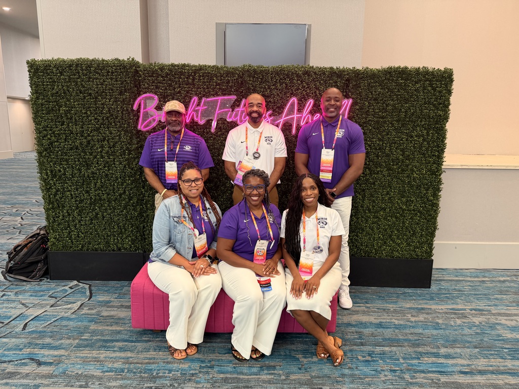 Representatives from Minor High School pose together for a photo at the Model Schools Conference. They stand and sit in front of a green background with neon pink letters attached to it.
