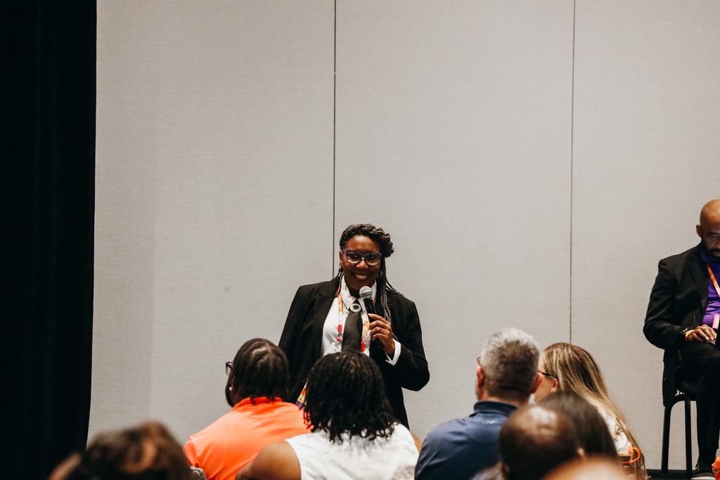 A woman smiles while speaking into a microphone at a conference. People sit at tables listening to her speak.