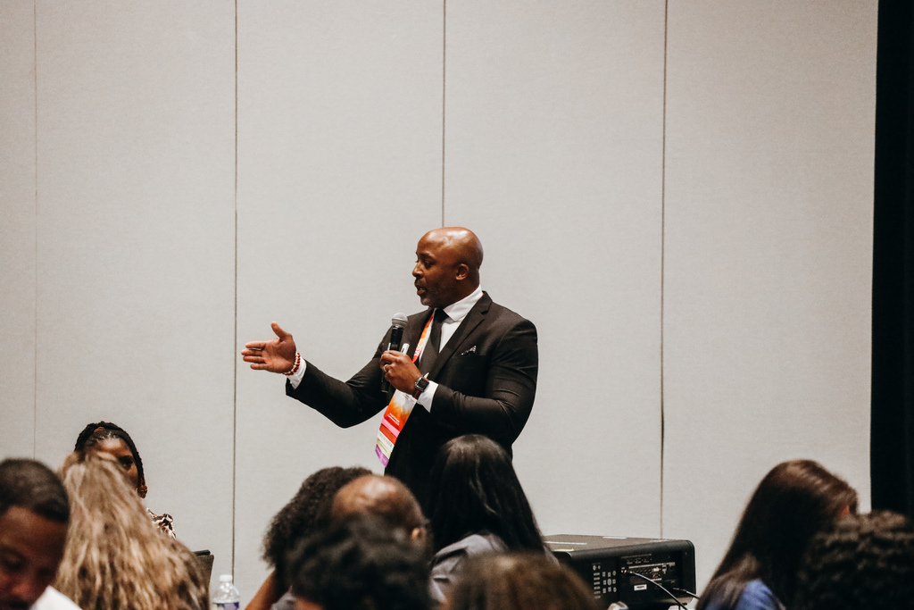 A man speaks into a microphone during a conference. People sit around him at tables listening.