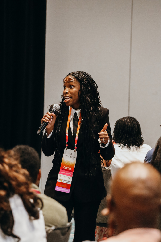 A woman speaks into a microphone during a conference. Educators sit around her at tables listening. The woman wears a black suit and tie.