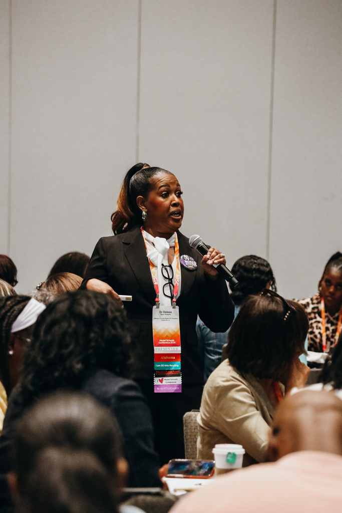 A woman speaks into a microphone at a conference. She wears a black blazer. Women sit around her at tables listening.
