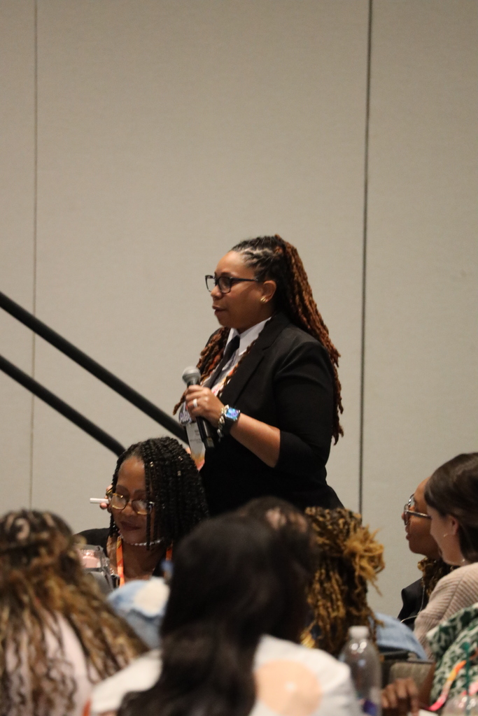 A woman speaks into a microphone while standing in a room. Women sit around her at tables.