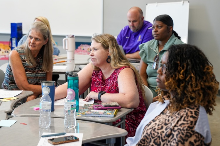 Educators and administrators sit at desks in a classroom and listen to speakers  