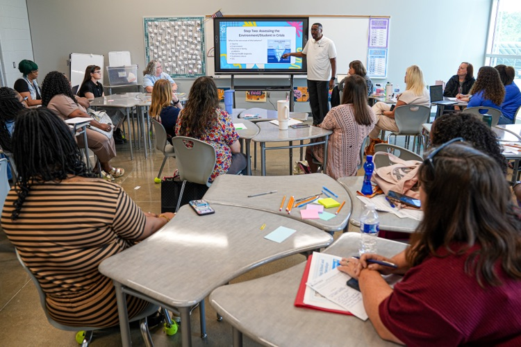 A man stands at a smart board and speaks to a group of educators and administrators as they sit at desks and listen  