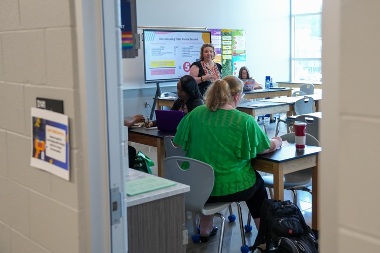 A woman stands at the front of a classroom and speaks to a group of educators and administrators as they sit at desks  