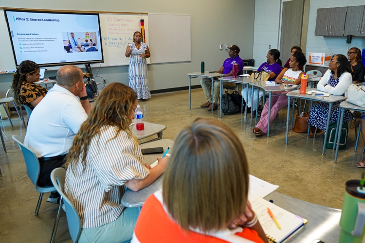 A woman stands at the front of a classroom and speaks to educators and administrators as they sit at desks  