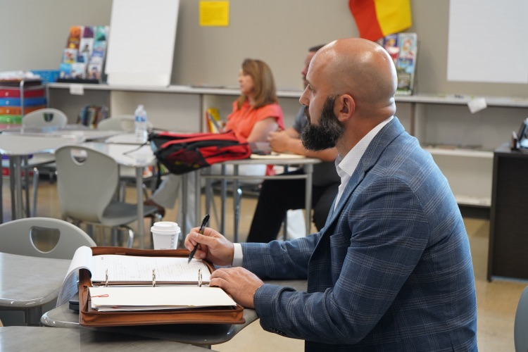 A man takes notes on paper while sitting at a desk in a classroom. 