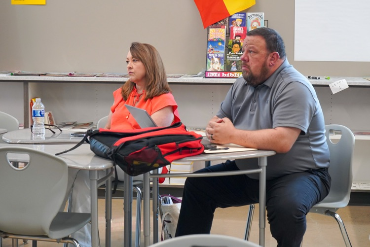 Administrators sit at desks in a classroom and listen to a speaker  